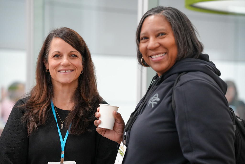 Two individuals smiling at a conference, one holding a coffee cup. The person on the right is wearing a hoodie with the GE (General Electric) logo.