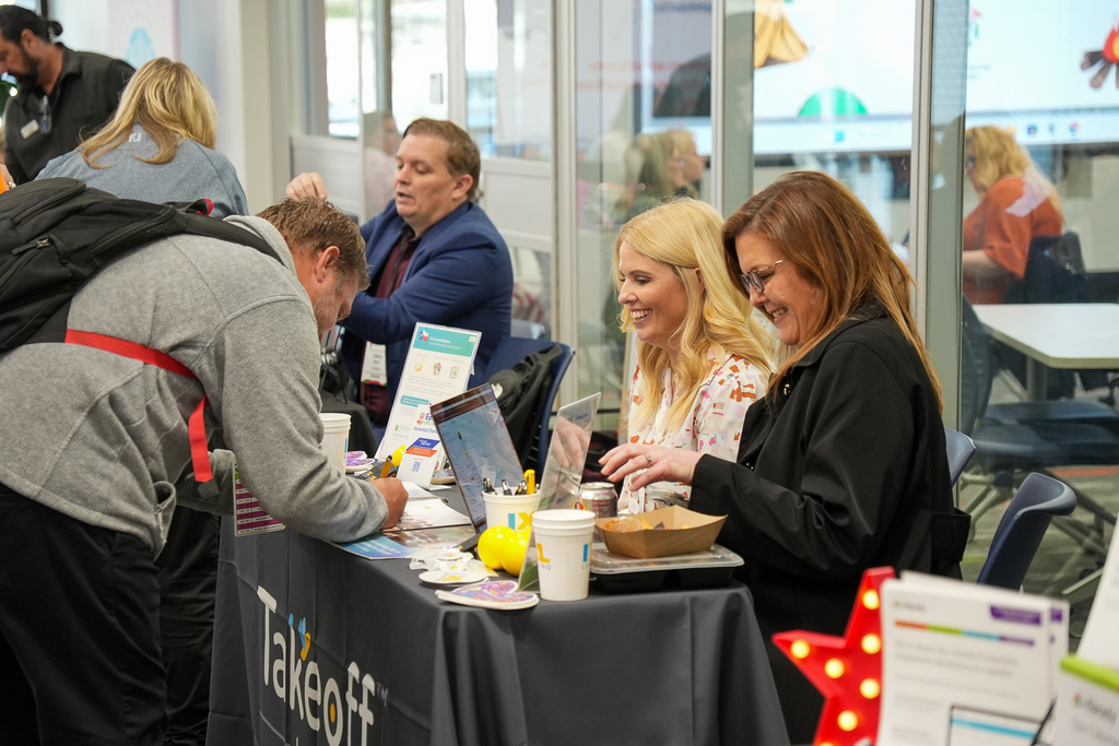 ttendees engage at a Takeoff conference booth, where people are discussing and exchanging information over a table with laptops and promotional materials.