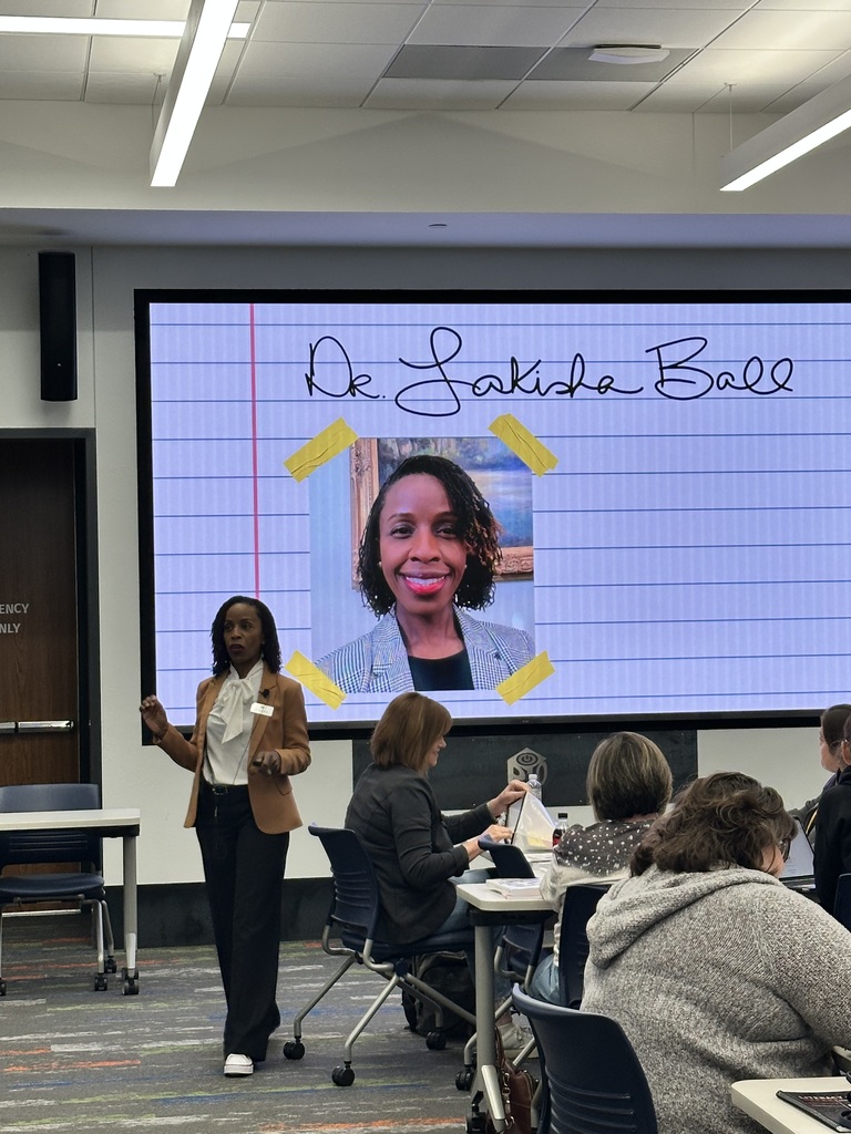 A speaker stands in front of an audience in a conference room. On a large screen behind, there is an image of a person and the text "Dr. Lakeida Ball." The audience is seated at tables, listening attentively.