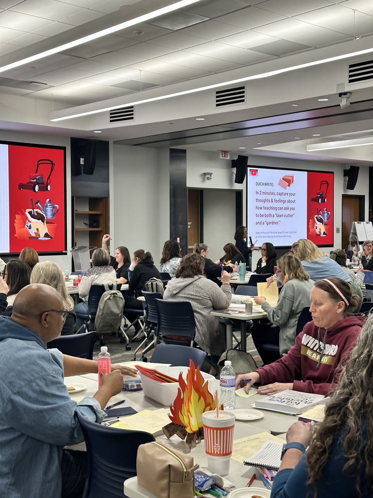 A group of people sitting at tables in a conference room, engaged in activities and discussions. There are presentations displayed on large screens at the front of the room.