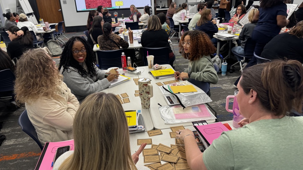 A group of people seated around tables in a classroom setting, engaging in discussions and activities. Various materials, such as papers and cards, are spread on the tables. The environment is busy and collaborative, with several people in the background also participating.