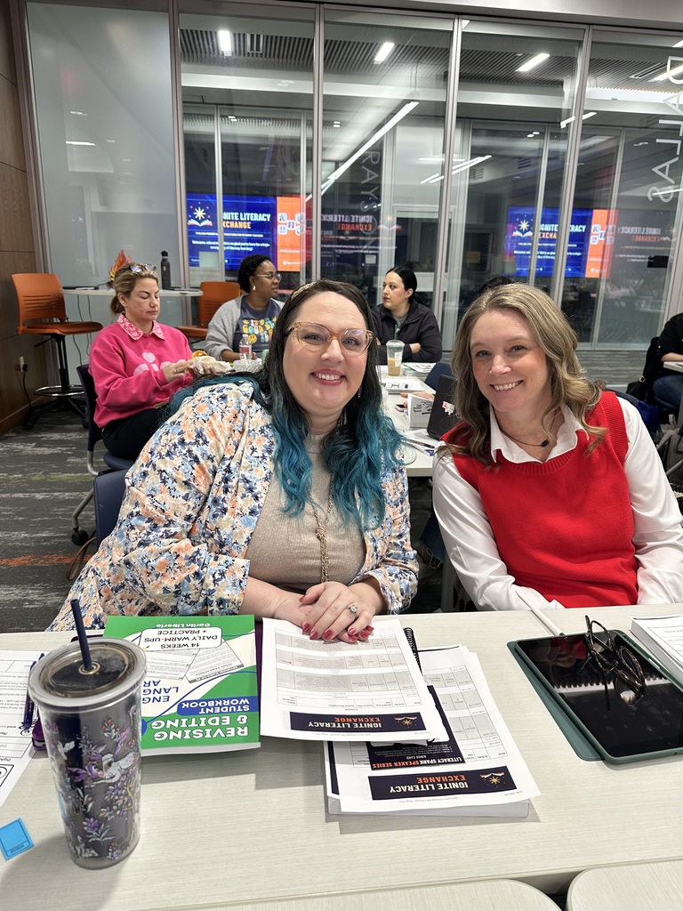 Two people sitting at a table in a study environment with books and papers. A water bottle is on the table. Other individuals are seated in the background, engaging with notebooks and screens. The setting appears to be a classroom or workshop.