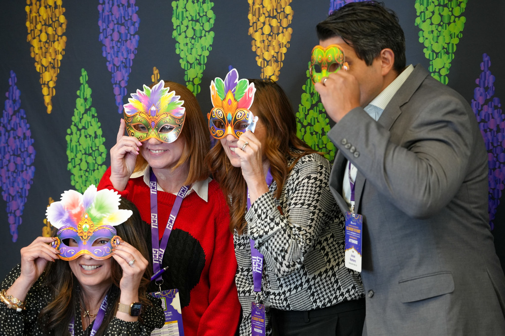 Four people stand in front of a festive backdrop with colorful cascading beads, holding decorative Mardi Gras masks over their faces. They wear conference badges, suggesting a formal event setting.