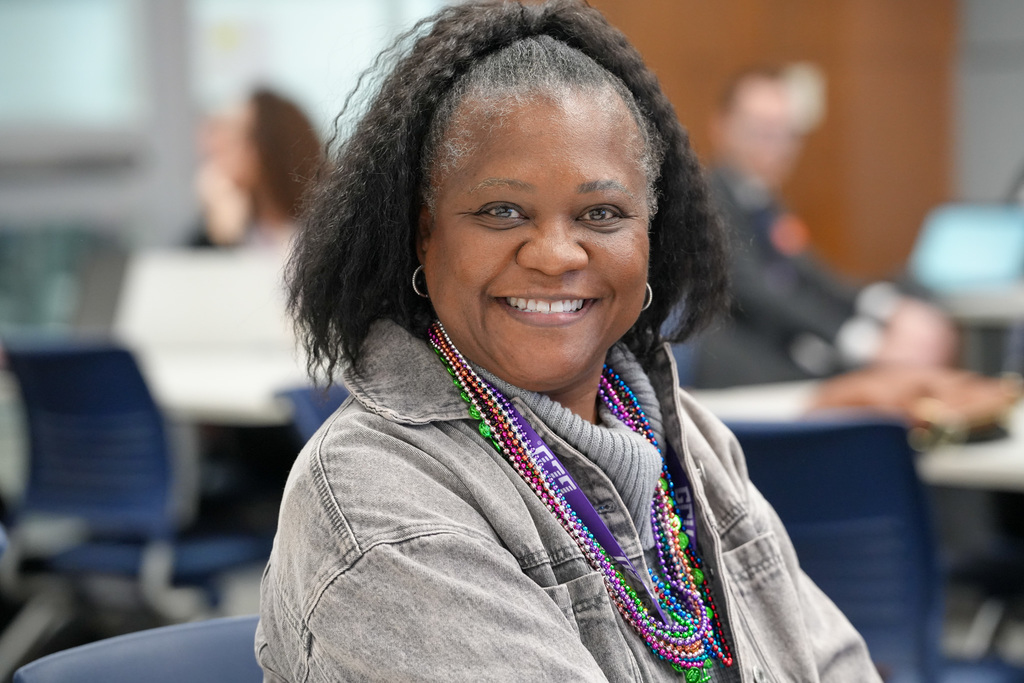 A smiling woman with shoulder-length black hair pulled half-up sits at a conference table, wearing a gray jacket and colorful bead necklaces. Blurred attendees and chairs are visible in the background, suggesting a professional meeting or training setting.