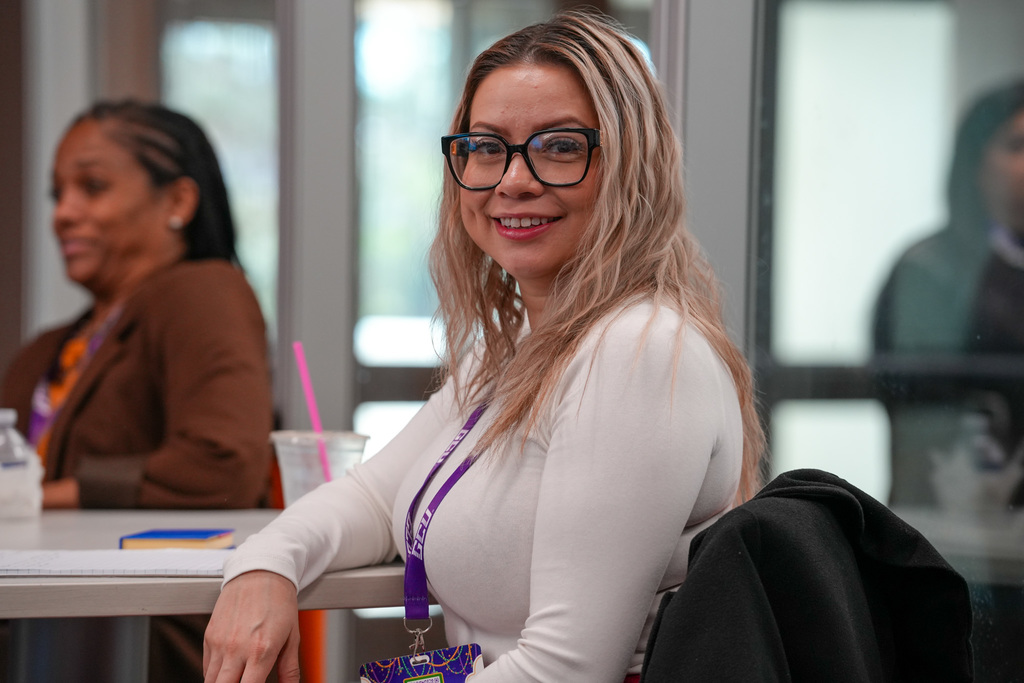 A woman with long blonde hair and black-framed glasses smiles while seated at a conference table. She wears a white top and a purple lanyard with a name badge. A notebook and drink cup with a pink straw sit on the table, and other attendees are visible in the blurred background.
