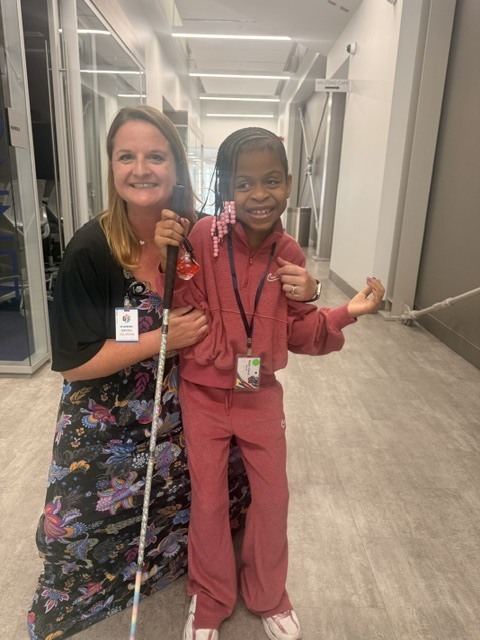 it shows two individuals smiling warmly, one holding a white cane, both dressed in colorful outfits, standing in a brightly lit hallway.