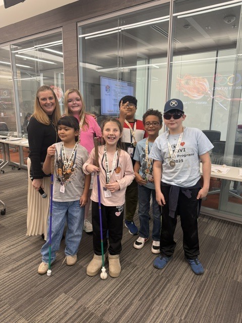 A group of seven people, including children and an adult, are standing together and smiling. The children are wearing lanyards with badges and holding canes. They are indoors, and behind them is a glass wall with the "AT&T" logo visible.