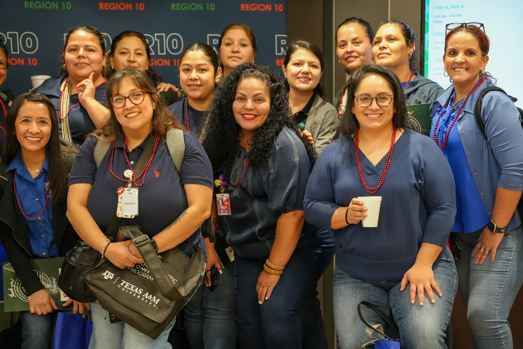 A group of women wearing navy tops and red bead necklaces pose together smiling in front of a “Region 10” backdrop, some holding bags and a cup, at what appears to be a conference or event.