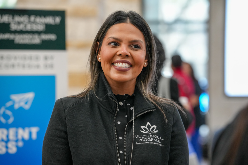 A person is smiling indoors at an event. They are wearing a dark jacket with "Multilingual Programs" and a logo on it. Blurred signage in the background reads "The Longhorn Family" and "Current Students."