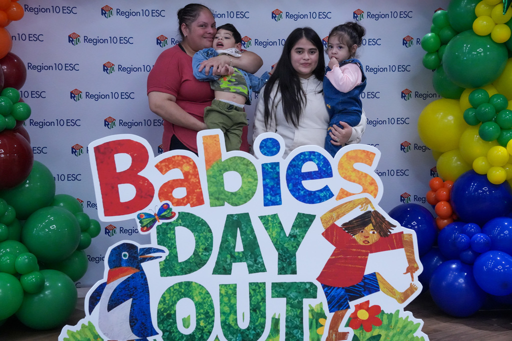 A group of people stands behind a colorful "Babies Day Out" sign, with vibrant balloon decorations on either side. A backdrop displays the "Region 10 ESC" logo repeatedly.