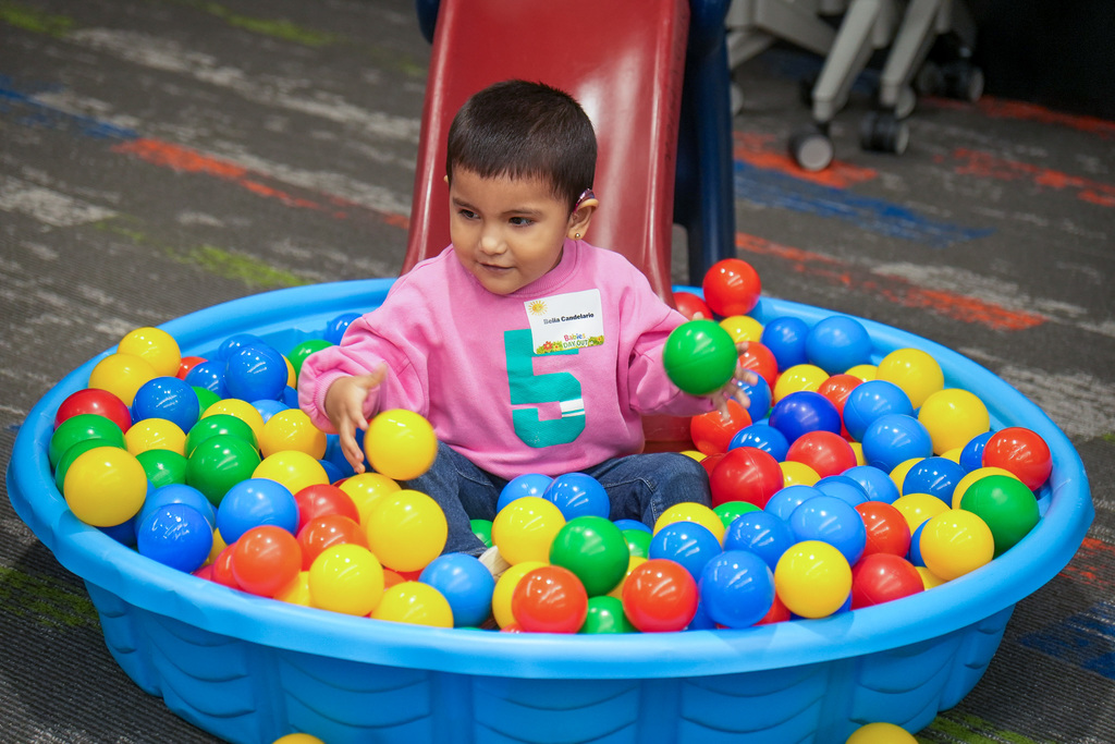 A young child sits in a blue plastic ball pit filled with colorful balls. The child is wearing a pink shirt with the number 5 printed on it. The background includes a carpeted floor with various colored patterns and a red slide.