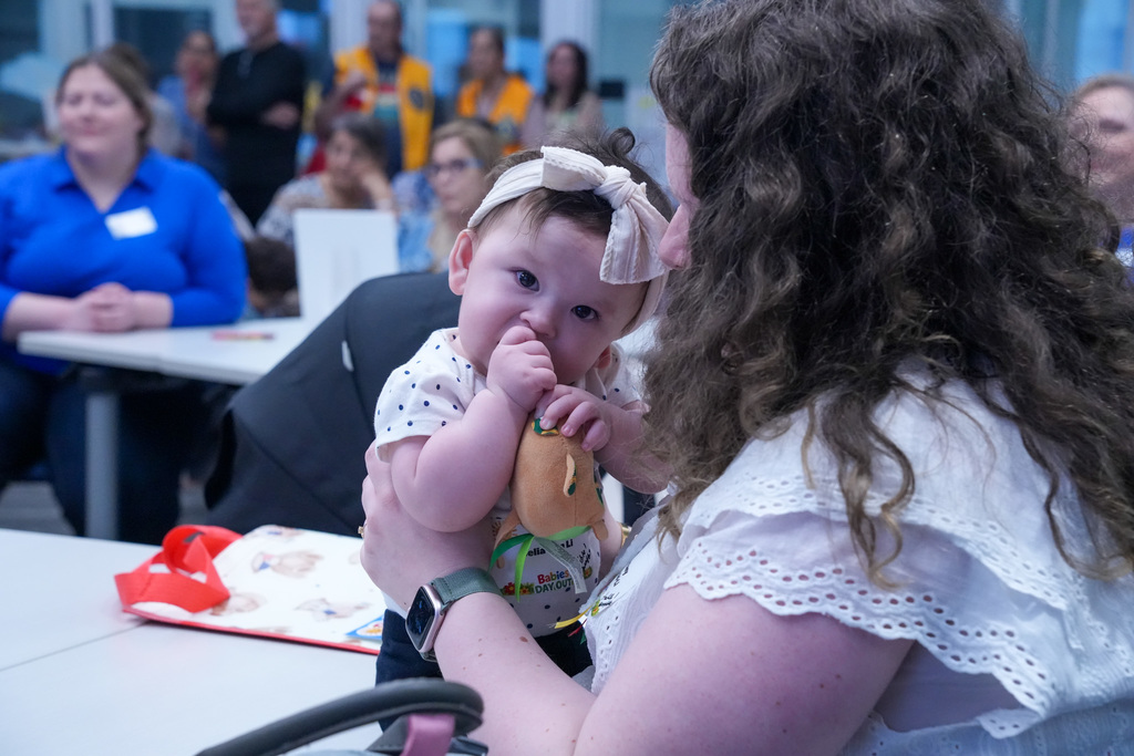 A person holding a baby who is chewing on a toy, surrounded by others seated in a classroom setting. There are bright colors in the background, and the atmosphere appears to be informal and relaxed.