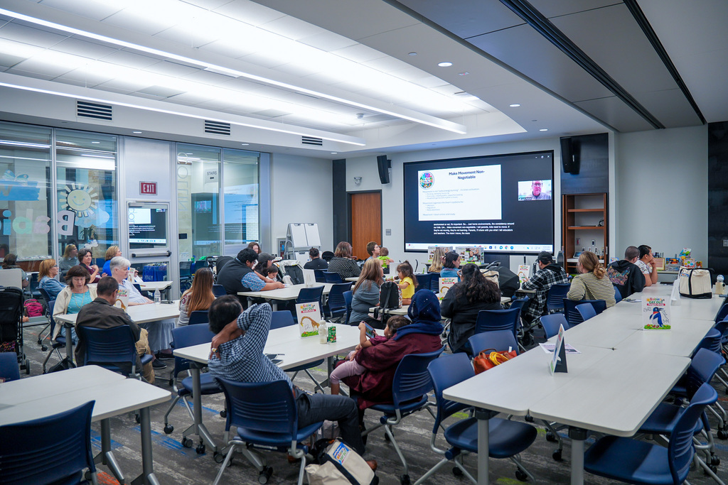 A group of people are seated in a conference room with modern furnishings. They are facing a large screen displaying a presentation, featuring a Windows logo. The room has whiteboards and glass partitions on one side. Tables have name placards and various items on them. The atmosphere is focused and collaborative.