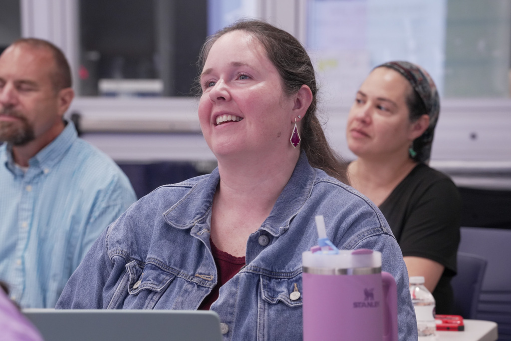 A person looking up front in a room full of people, smiling at the presenter.