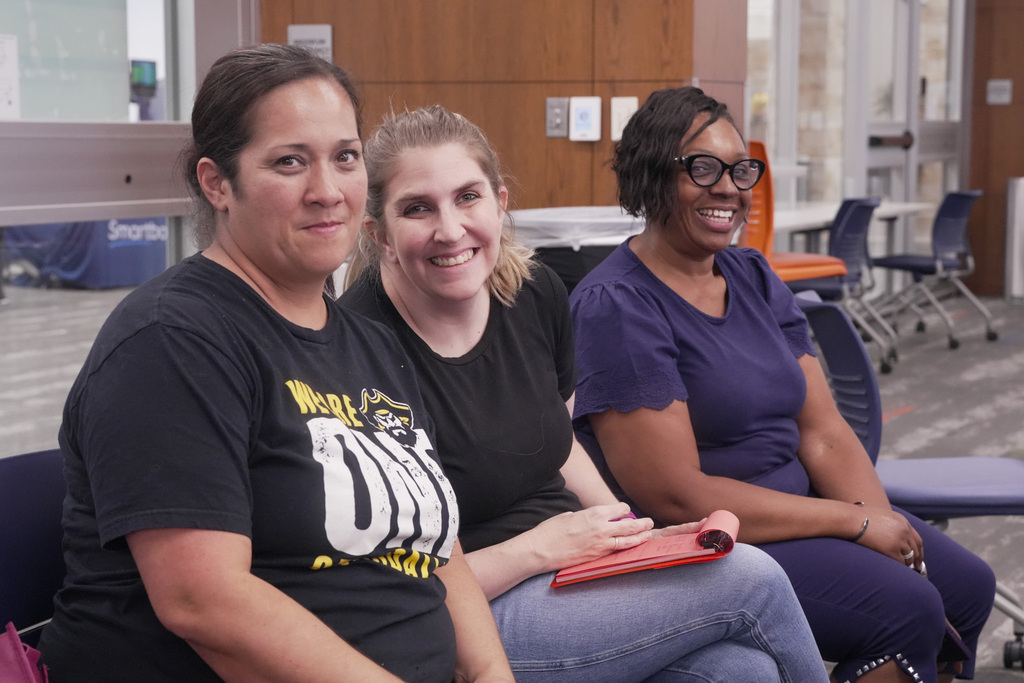 Three individuals sitting together indoors, smiling. The person on the left is wearing a black t-shirt, the one in the middle is in a light top holding a red item, and the person on the right is wearing glasses and a dark top. They are seated in a room with tables and chairs.