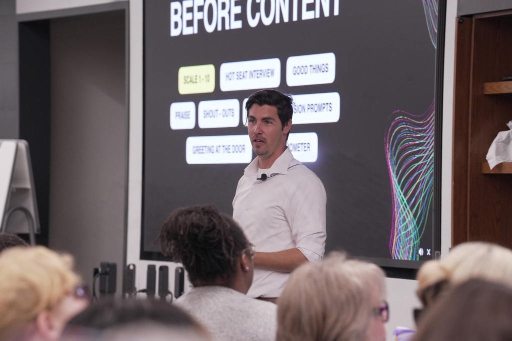  person stands in front of a seated audience, presenting near a screen displaying the text "BEFORE CONTENT." The screen lists phrases such as "SCALE 1-10," "HOT SEAT INTERVIEW," and "GOOD THINGS." There is a colorful abstract pattern on the right side of the screen.