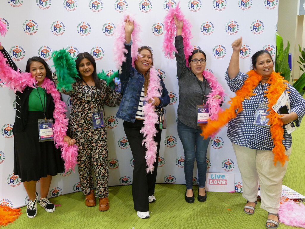A group of five people is smiling and standing in front of a backdrop with repeating logos. They are celebrating with colorful feather boas in pink, green, and orange. Some are holding up their arms enthusiastically. They are wearing conference badges and standing on green carpet, with a small sign reading "Live Love" on the floor.