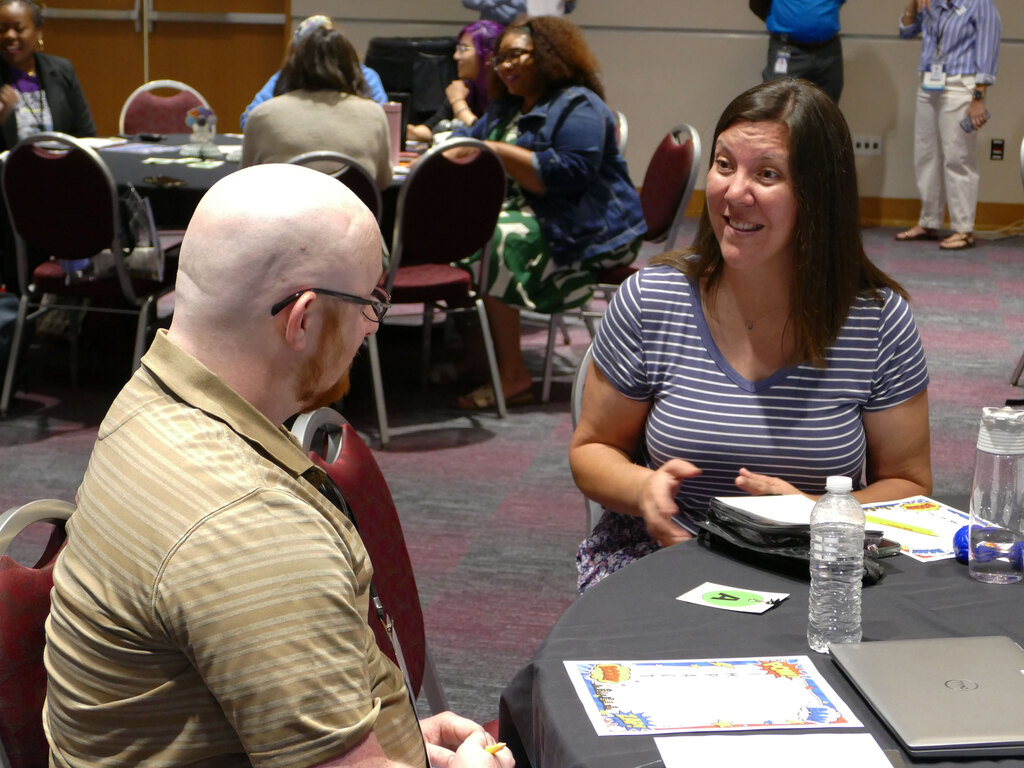 A person with glasses is seated at a round table, facing another person who is smiling and holding a notebook. The table has a certificate and a water bottle on it. In the background, other people are gathered around similar tables in a conference room setting.