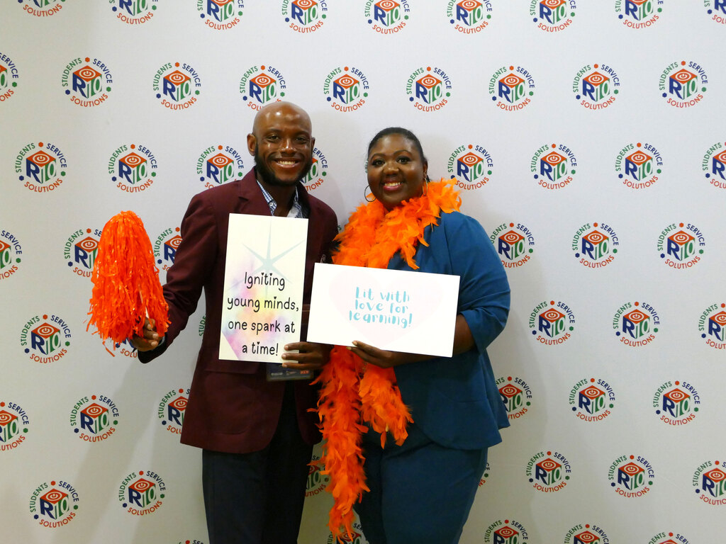 Two people smiling and holding signs in front of a background featuring repeated Rio Ferdinand Foundation logos. One person holds an orange pom-pom and a sign that reads, "Igniting young minds, one spark at a time!" The other holds a sign that says, "Lit with love for learning!" Both wear orange feather boas.