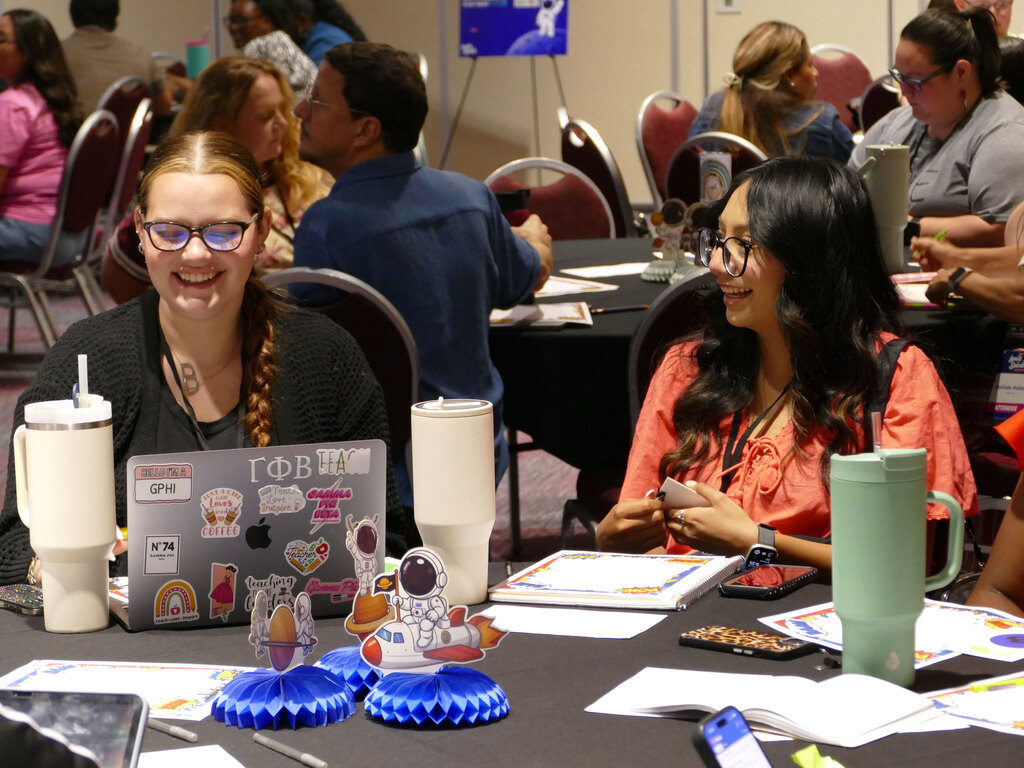 A group of people seated around a table at a conference. Two individuals are in focus, smiling and engaged in conversation. The table has laptops adorned with various stickers, including an astronaut and rainbows. Other attendees are in the background, also interacting. The setting is casual and collaborative.