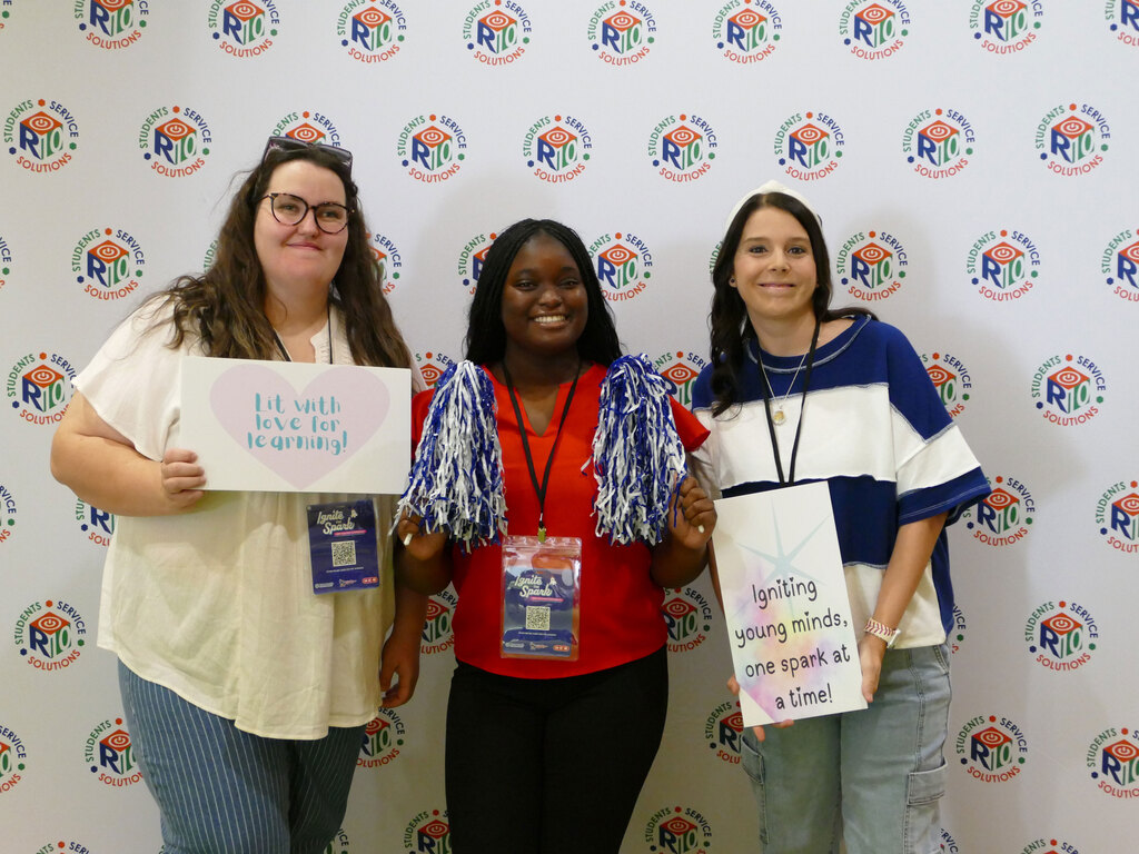 Three individuals are standing in front of a backdrop with "Rio" Foundation logos. They are smiling and holding signs that say, "Lit with love for learning!" and "Igniting young minds, one spark at a time!" Each person is wearing a lanyard with a badge.