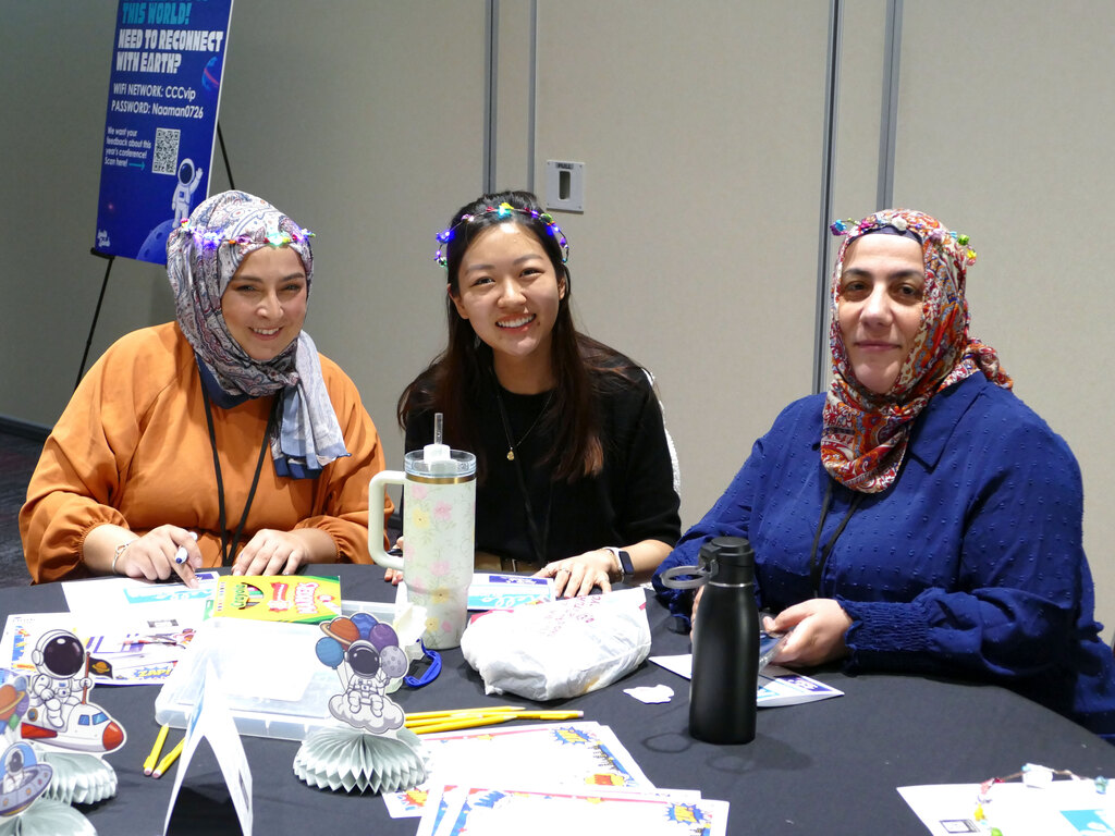 Three people are seated at a table, smiling and working on crafts. The table is filled with colorful materials, including paper and drawings of NASA-related themes. A sign in the background mentions NASA.