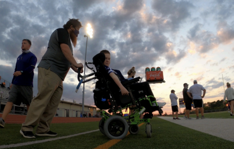 Jarren McTee sideline at a football game working in his wheelchair and with a teacher