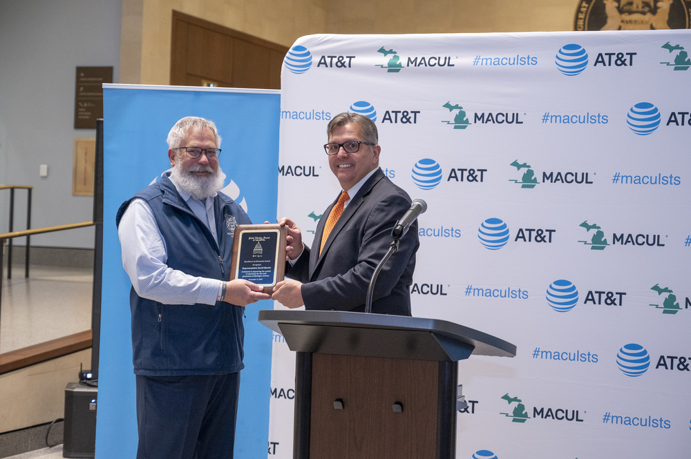  Photos from the Student Technology Showcase at the Michigan State Capitol feature an award presentation, a large check presentation, and students demonstrating their technology projects. In the first image, two men stand in front of an AT&T and MACUL backdrop as one receives a plaque. In the second, a man and woman hold an oversized check awarded to MACUL. The third image shows students and adults gathered around a table display featuring digital and hands-on projects created by Manchester Schools.