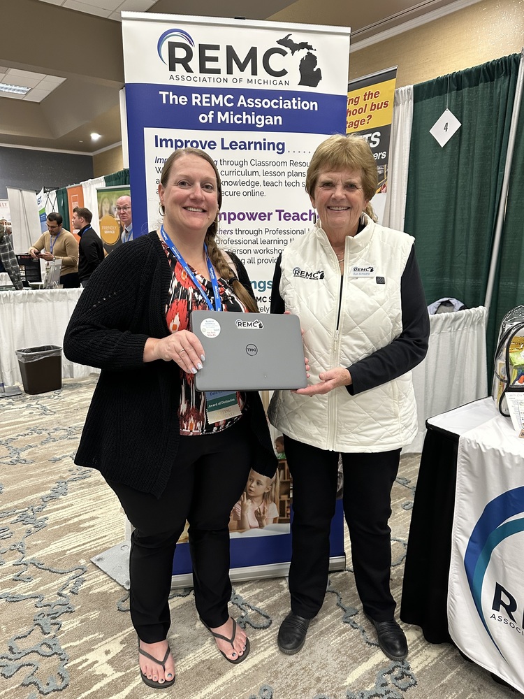 Two women stand smiling in front of a REMC Association of Michigan booth at the MASB conference. Both women are smiling while one holds a silver Dell laptop awarded as a prize from REMC SAVE. Behind them is a standing banner with REMC branding and the phrases “Improve Learning” and “Empower Teachers.” The booth is set up with other tables and attendees visible in the background.