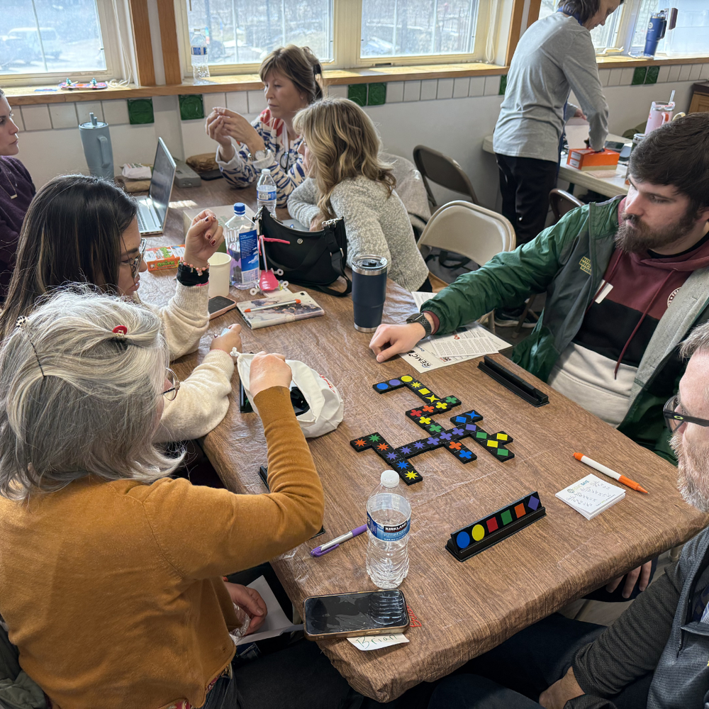  group of adults sit around a table indoors at Leslie Science & Nature Center, collaborating during a STEM in Action Day event focused on AI for the classroom. Participants are engaged in hands-on activities using colorful pattern blocks and game-like materials spread across the table, with laptops, notebooks, and water bottles nearby. The setting is bright with large windows in the background, and individuals appear focused and engaged in discussion and exploration of STEM teaching tools.