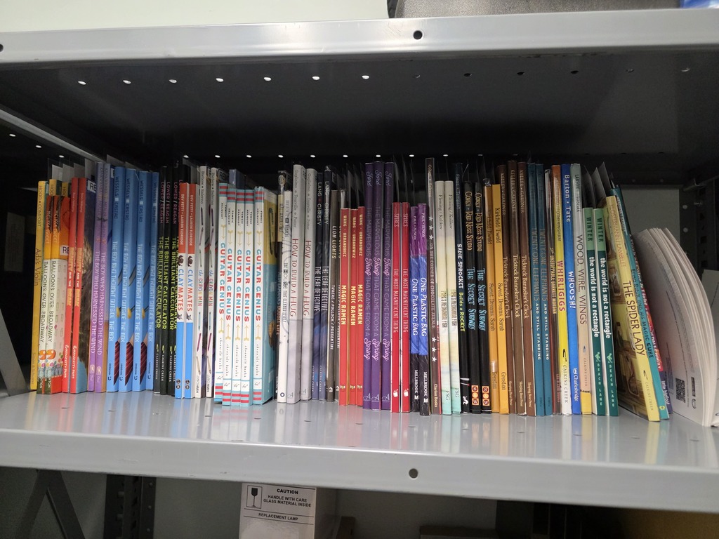 A front-facing view of a light grey metal shelf filled with a neat row of slim children's picture books and non-fiction titles. The spines feature vibrant colors including red, blue, yellow, and white, with visible titles such as Balloons Over Broadway, Guitar Genius, Magic Ramen, and The Spider Lady. On the far right, a few softcover books and a thick white volume lean against the end panel. A small sticker with a caution warning is affixed to the shelf rim underneath.