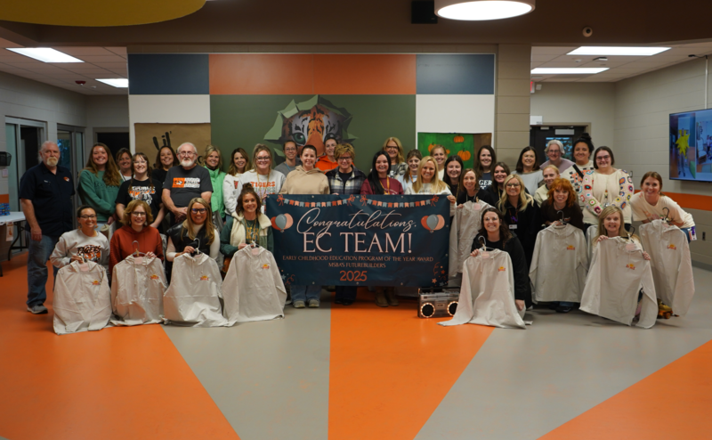 Early Childhood Center staff standing together and smiling, while holding a banner celebrating their award and jackets
