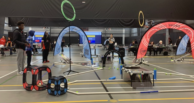 “A wide view of a school gym during a drone competition with teams, spectators, and competition fields set up on the floor.”