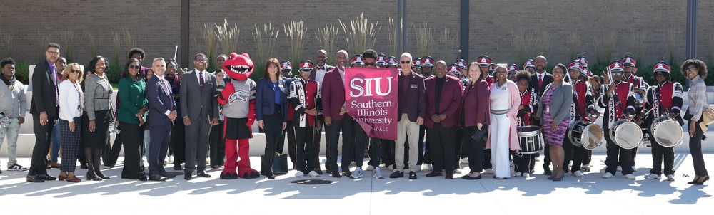 Group photo outdoors with school and university leaders, students, the marching band, and a red mascot; two people hold an “SIU Southern Illinois University” banner.