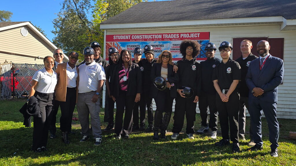 English: Students, staff, and project partners gather outside the Rich Township Construction Trades project house. Several students wear black polos and hold or wear hard hats. A sign behind the group reads “Student Construction Trades Project: Real Homes. Real Skills. Real Impact.”  Español: Estudiantes, personal y colaboradores se reúnen frente a la vivienda del proyecto de Oficios de la Construcción de Rich Township. Varios estudiantes llevan polos negros y sostienen o usan cascos. Detrás del grupo, un letrero dice: “Proyecto estudiantil de construcción: viviendas reales, habilidades reales, impacto real”.