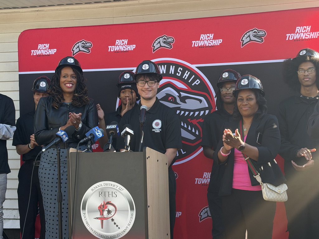 A Rich Township construction trades student wearing a black polo and hard hat smiles behind a podium with news microphones as fellow students and adults applaud. A red and black Rich Township backdrop frames the home project announcement. Un estudiante del programa de Oficios de la Construcción de Rich Township, con polo negro y casco, sonríe detrás de un podio con micrófonos de medios de comunicación mientras otros estudiantes y adultos aplauden. Un fondo rojo y negro de Rich Township enmarca el anuncio del proyecto de renovación de una vivienda.