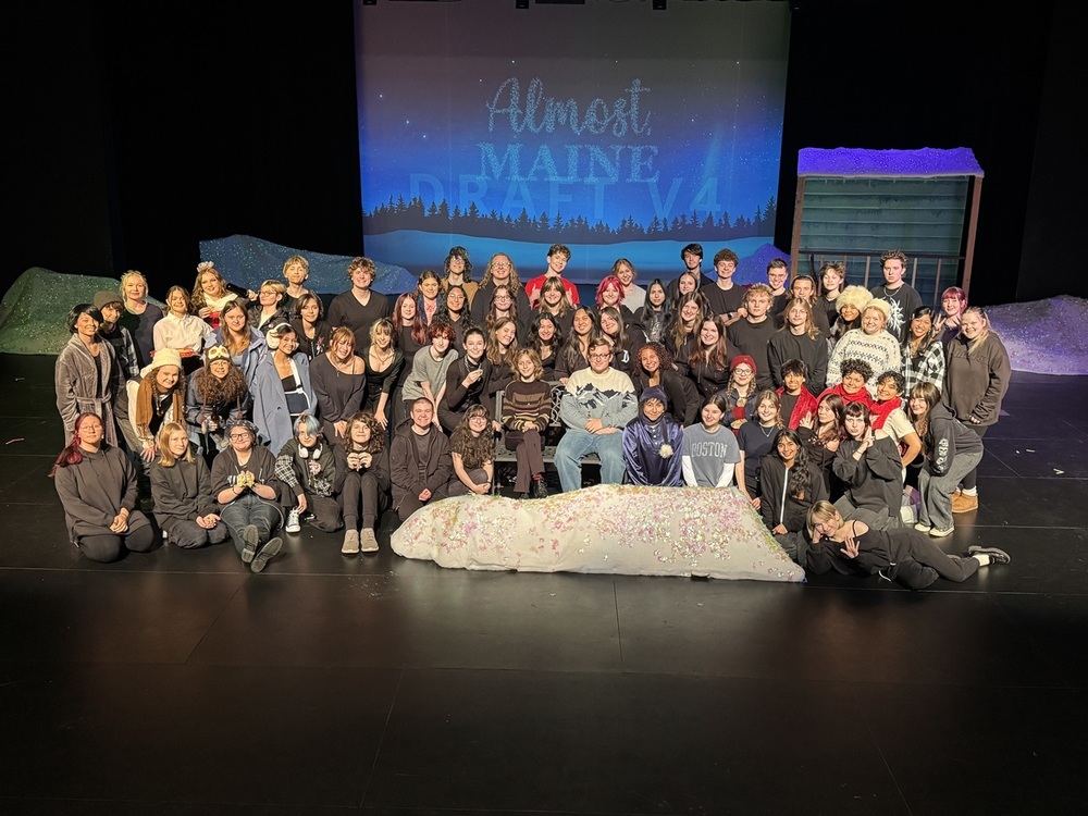 The entire cast and crew posing on stage in front of the projector screen with "Almost, Maine" projected on the screen