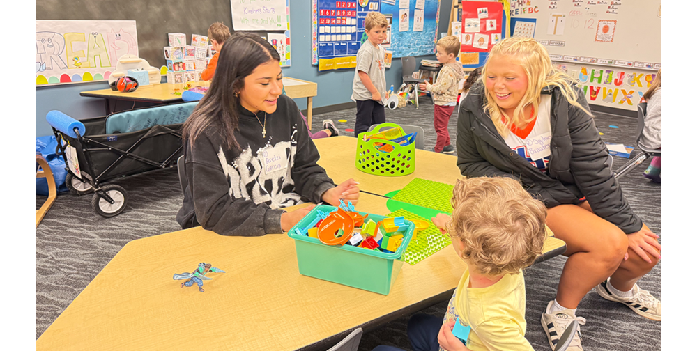 High school  students sit at a table with a preschool student who is playing with colorful toys