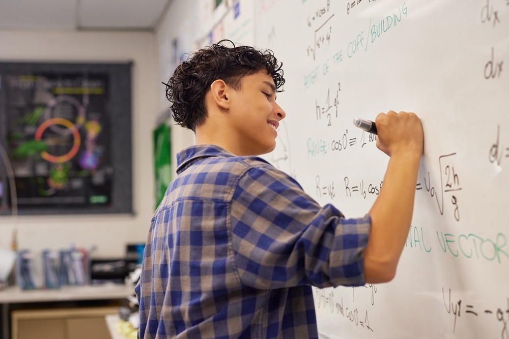 A male high school student smiling while working through a math problem on a whiteboard