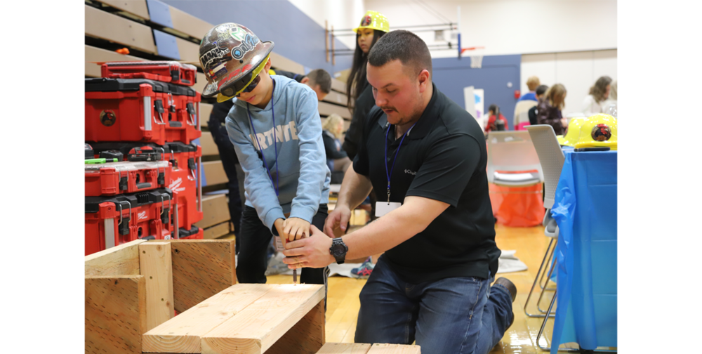 An adult male carpenter helps a 4th grade student insert a screw into wooden staircase
