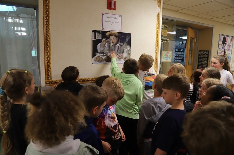 a group of about 20 elementary school students in the hallway looking at an art print of a man eating beans. Above the artwork is a sign that reads "How does food nourish us?"