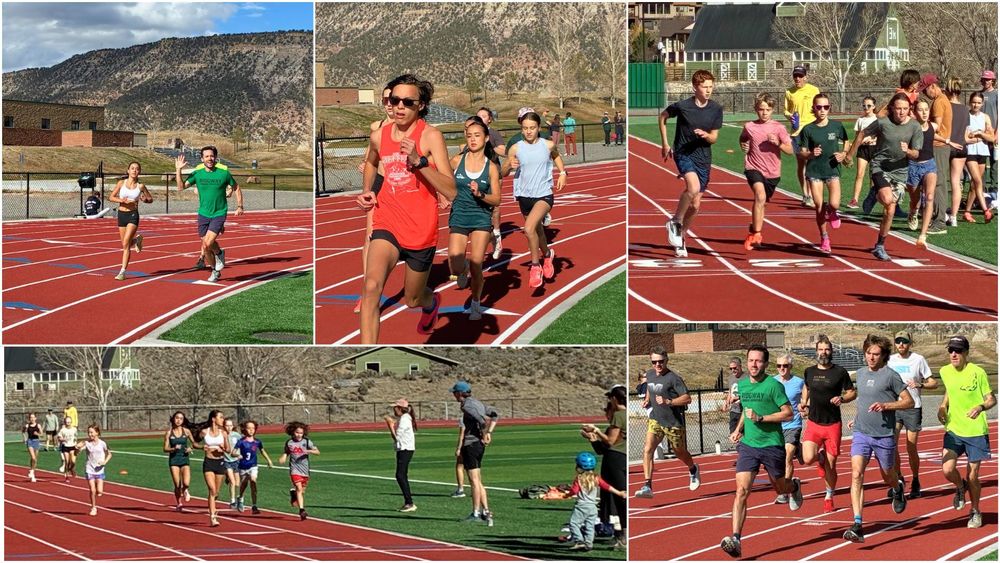 A group of children and teens run together on a red track during a community event, with several adults standing on the infield and sidelines cheering them on. The scene takes place on a sunny day with grassy fields and buildings visible in the background.