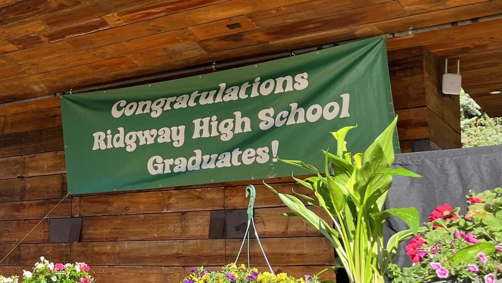 Banner reading “Congratulations Ridgway High School Graduates!” displayed above flowers and plants at an outdoor venue.