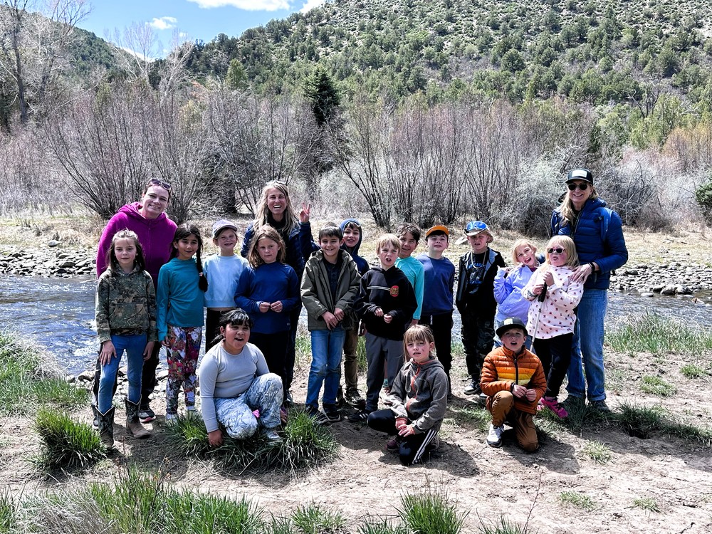 Group of second grade students and adults standing by a creek at Pa-Co-Chu-Puk, smiling outdoors during a field learning experience.