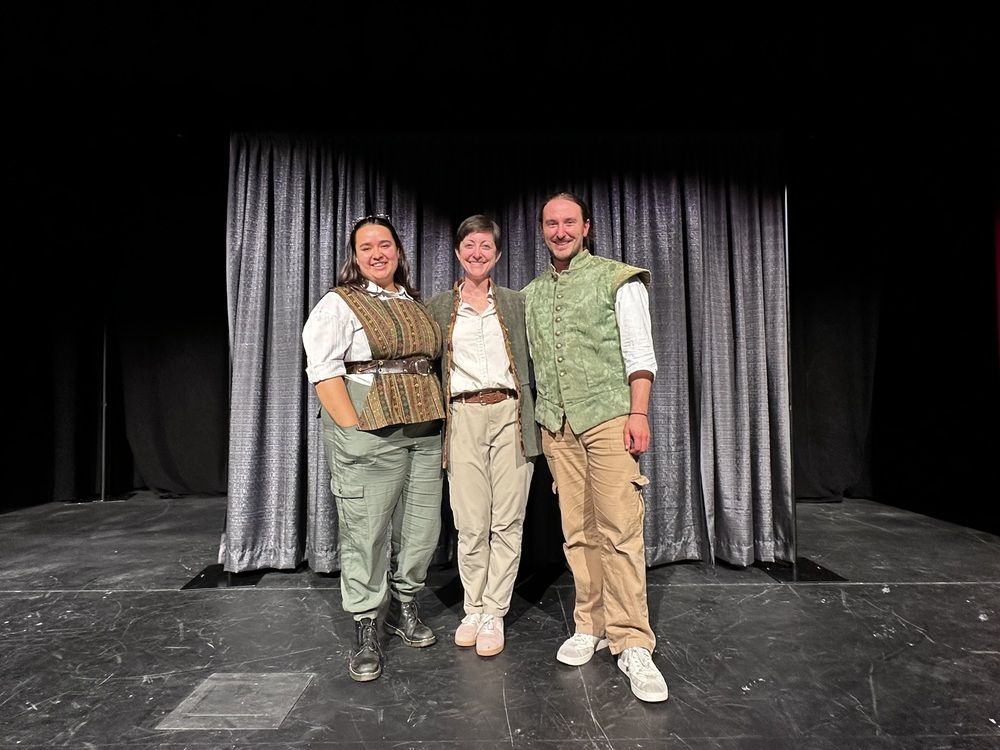 Three people stand smiling on a stage in front of a black curtain, wearing theatrical costumes after a performance.
