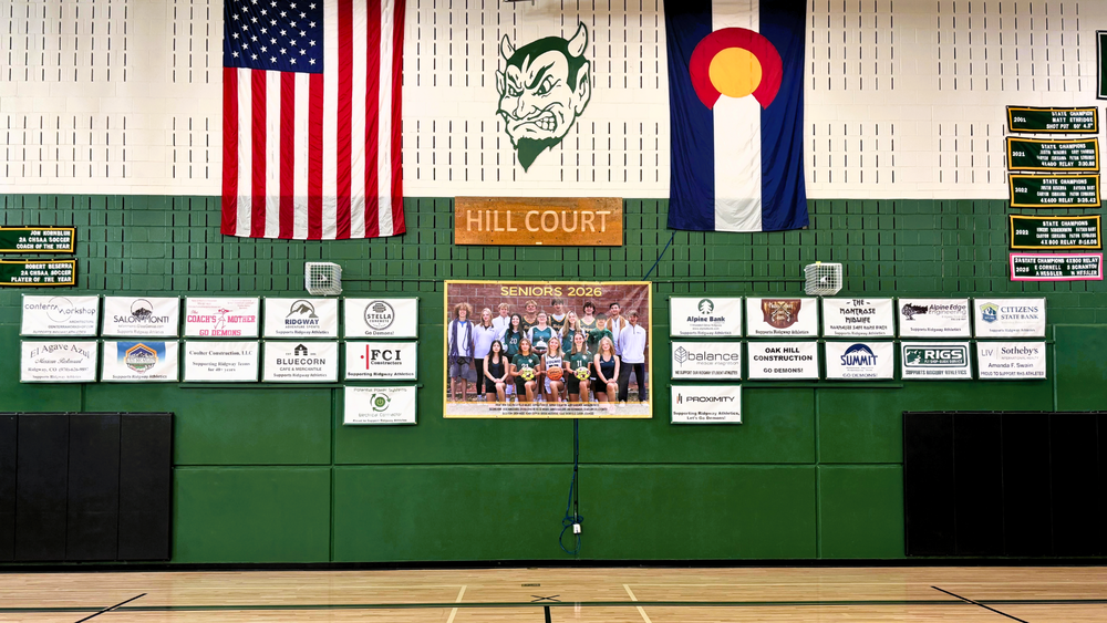 Gymnasium wall with U.S. and Colorado flags, a large demon mascot logo, and sponsor banners displayed in rows. A large team photo labeled ‘Seniors 2026’ is centered among the banners. Green padded walls and a wooden gym floor are visible.