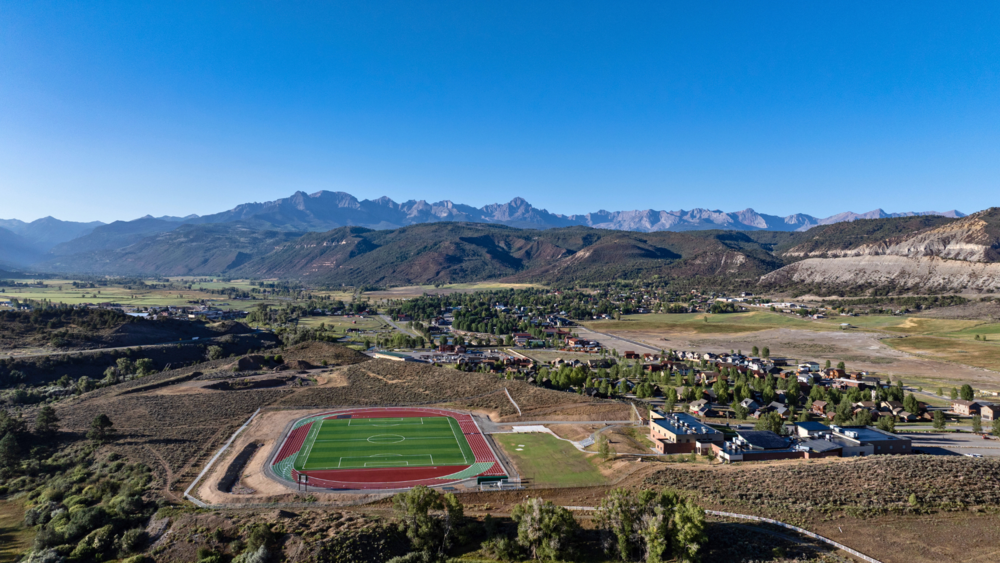 Aerial view of the Cimarron Athletic Field with track, town, and mountains in the background under a clear blue sky.