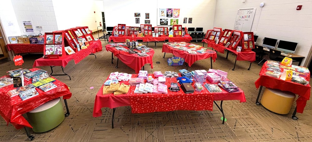 School library book fair setup with tables of children’s books and small gifts displayed on red tablecloths in a large room.