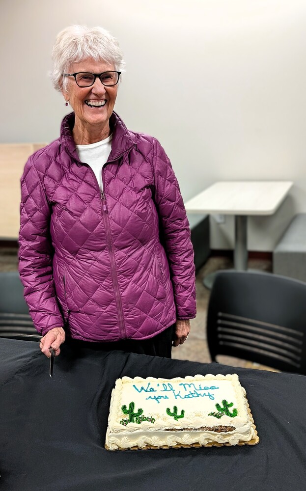 Smiling woman in a purple jacket stands behind a farewell cake that reads, “We’ll miss you Kathy.”
