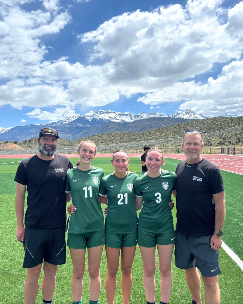 Three High School girls soccer seniors stand with their coaches on the field, with mountain views and blue skies in the background.